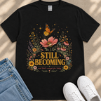 a black t-shirt with a floral design and the text "STILL BECOMING" printed on it, along with a pair of jeans and white sneakers.