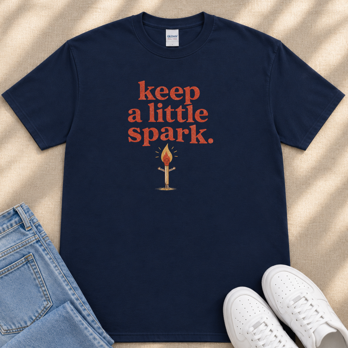 A navy blue t-shirt with the text "keep a little spark" printed on it, along with an illustration of a lit match. The t-shirt is placed next to a pair of blue jeans and white sneakers.