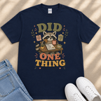 A navy blue t-shirt with a graphic design featuring a raccoon holding a sign that says "Did One Thing" and surrounded by various objects, such as a cup, a book, and a plant.