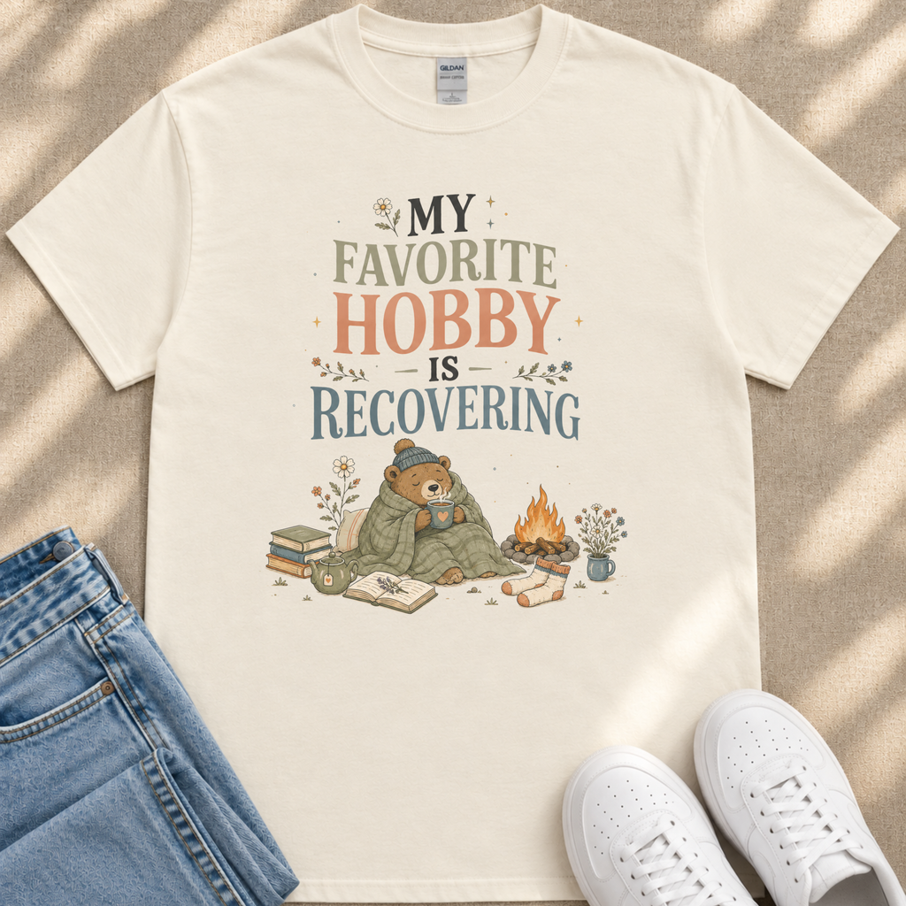 A white t-shirt with a printed design of a bear wrapped in a blanket, sitting on a blanket and reading a book, with a campfire and flowers in the background.