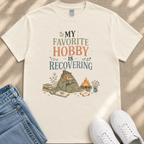 A white t-shirt with a printed design of a bear wrapped in a blanket, sitting on a blanket and reading a book, with a campfire and flowers in the background.