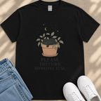 A black t-shirt with a graphic design depicting a cat sleeping in a pot, accompanied by the text "PLEASE DISTURB SOMEONE ELSE." The t-shirt is placed on a beige surface, with a pair of blue jeans and white sneakers visible in the background.