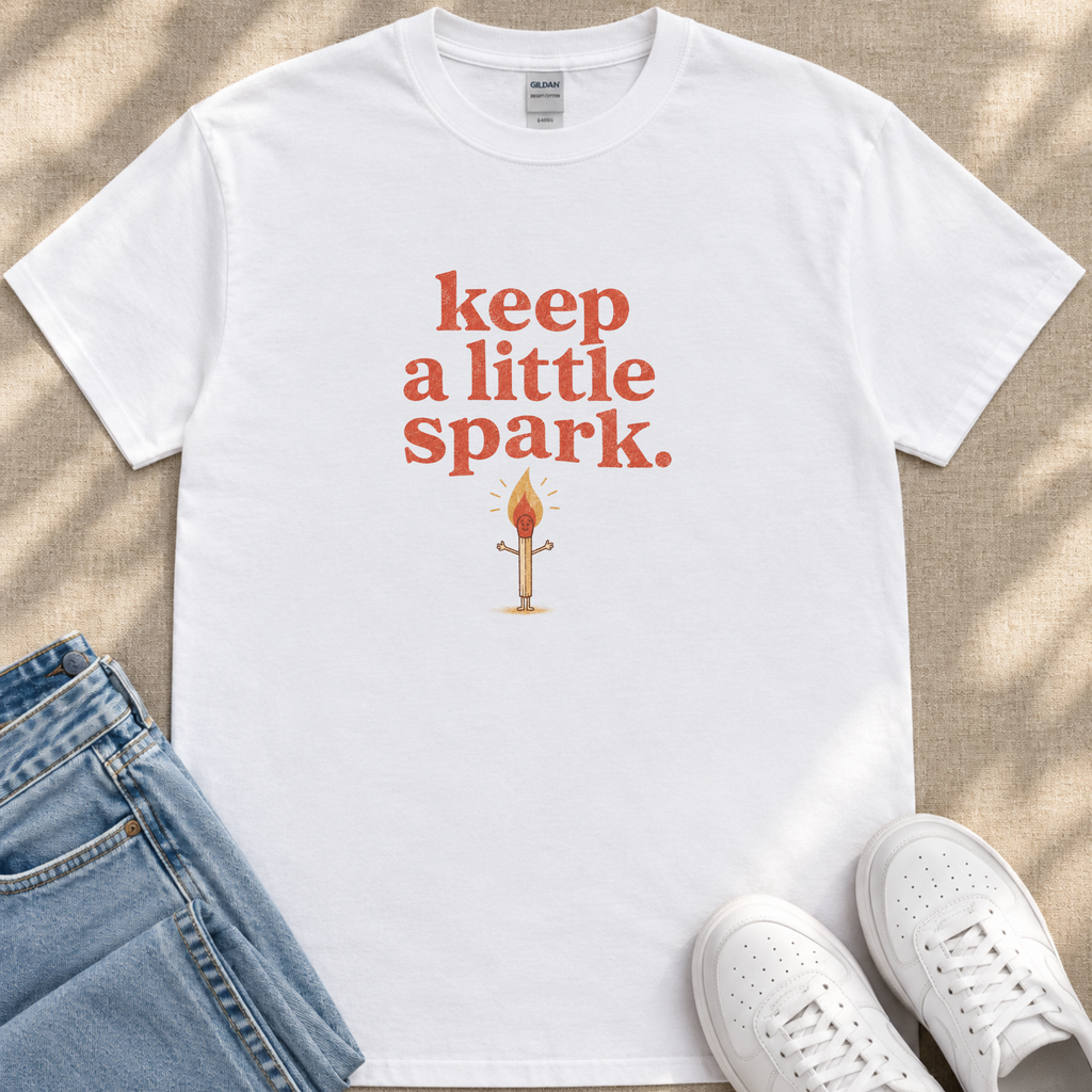 A white t-shirt with the text "keep a little spark" printed on it, along with an illustration of a lit match. The t-shirt is placed on a beige surface, and there are a pair of blue jeans and white sneakers visible in the image.
