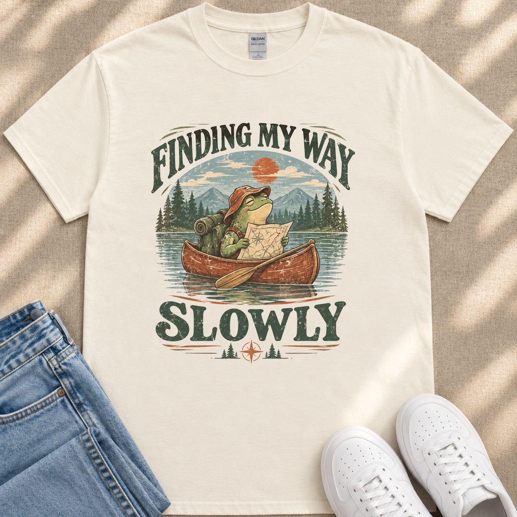 A white t-shirt with a graphic design depicting a frog in a canoe, holding a map and paddling slowly, with the text "FINDING MY WAY SLOWLY" printed on it.