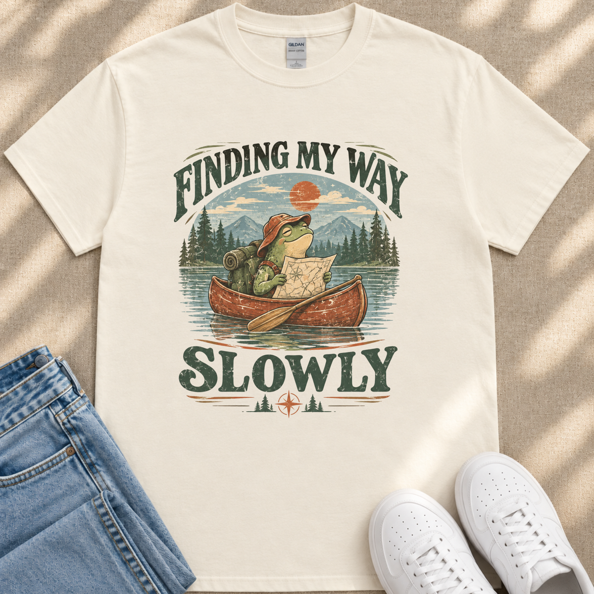 A white t-shirt with a graphic design depicting a frog in a canoe, holding a map and paddling slowly, with the text "FINDING MY WAY SLOWLY" printed on it.