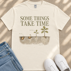 A white t-shirt with a graphic design depicting the growth of a plant, along with the text "Some things take time" and illustrations of the plant's first leaves, roots, and a seed.