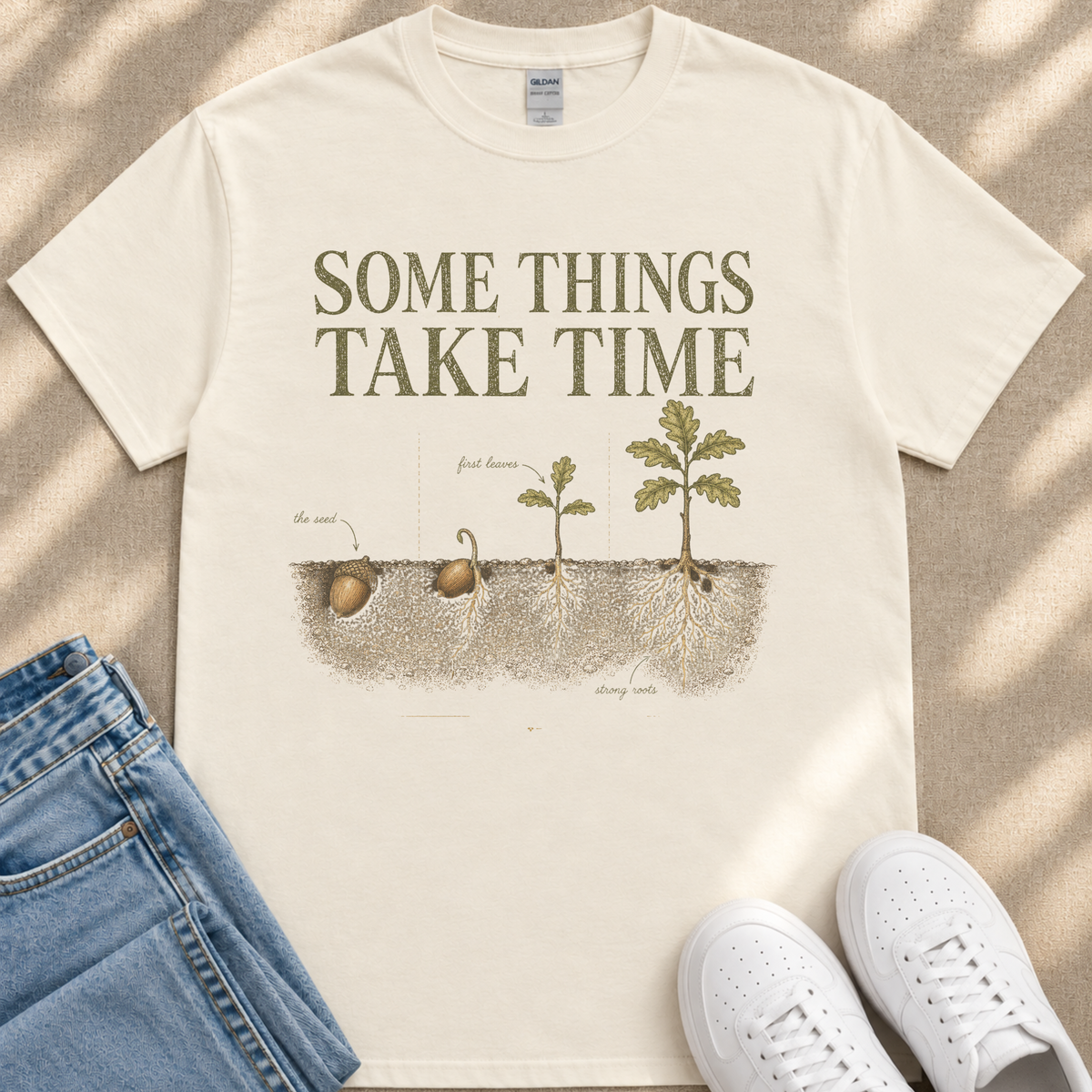 A white t-shirt with a graphic design depicting the growth of a plant, along with the text "Some things take time" and illustrations of the plant's first leaves, roots, and a seed.