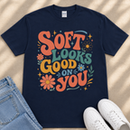 A navy blue t-shirt with a colorful graphic design featuring the text "Soft looks good on you" surrounded by flowers and stars.