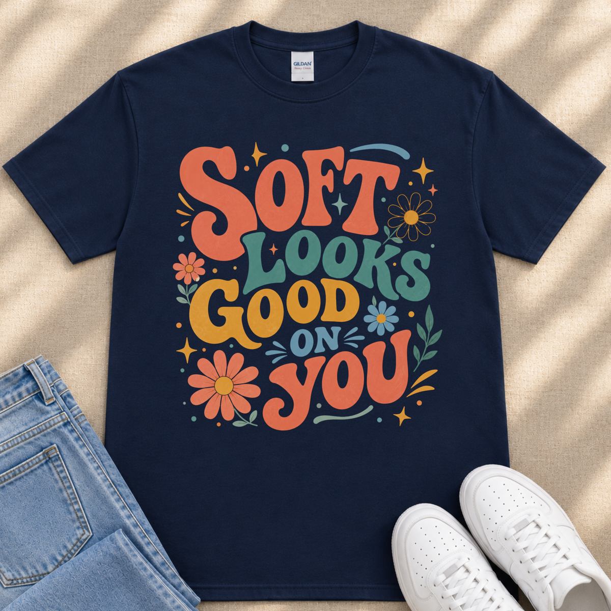 A navy blue t-shirt with a colorful graphic design featuring the text "Soft looks good on you" surrounded by flowers and stars.