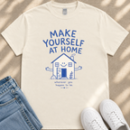 A white t-shirt with a blue graphic design and text, along with a pair of jeans and white sneakers, laid out on a beige surface.