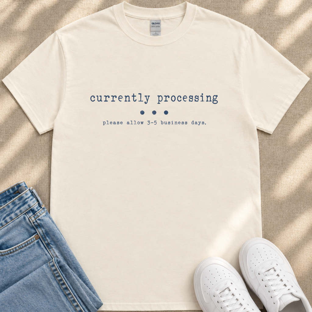 A white t-shirt with the text "currently processing" printed on it, along with a message that says "please allow 3-5 business days." The t-shirt is laid out on a beige surface, and there are a pair of jeans and white sneakers visible in the image.
