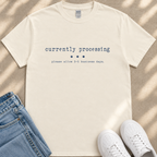 A white t-shirt with the text "currently processing" printed on it, along with a message that says "please allow 3-5 business days." The t-shirt is laid out on a beige surface, and there are a pair of jeans and white sneakers visible in the image.
