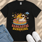 A black t-shirt with a colorful graphic design depicting a duck wearing sunglasses and sitting in a pool, surrounded by sun and clouds, along with the text "Your little ray of SARCASTIC SUNSHINE".