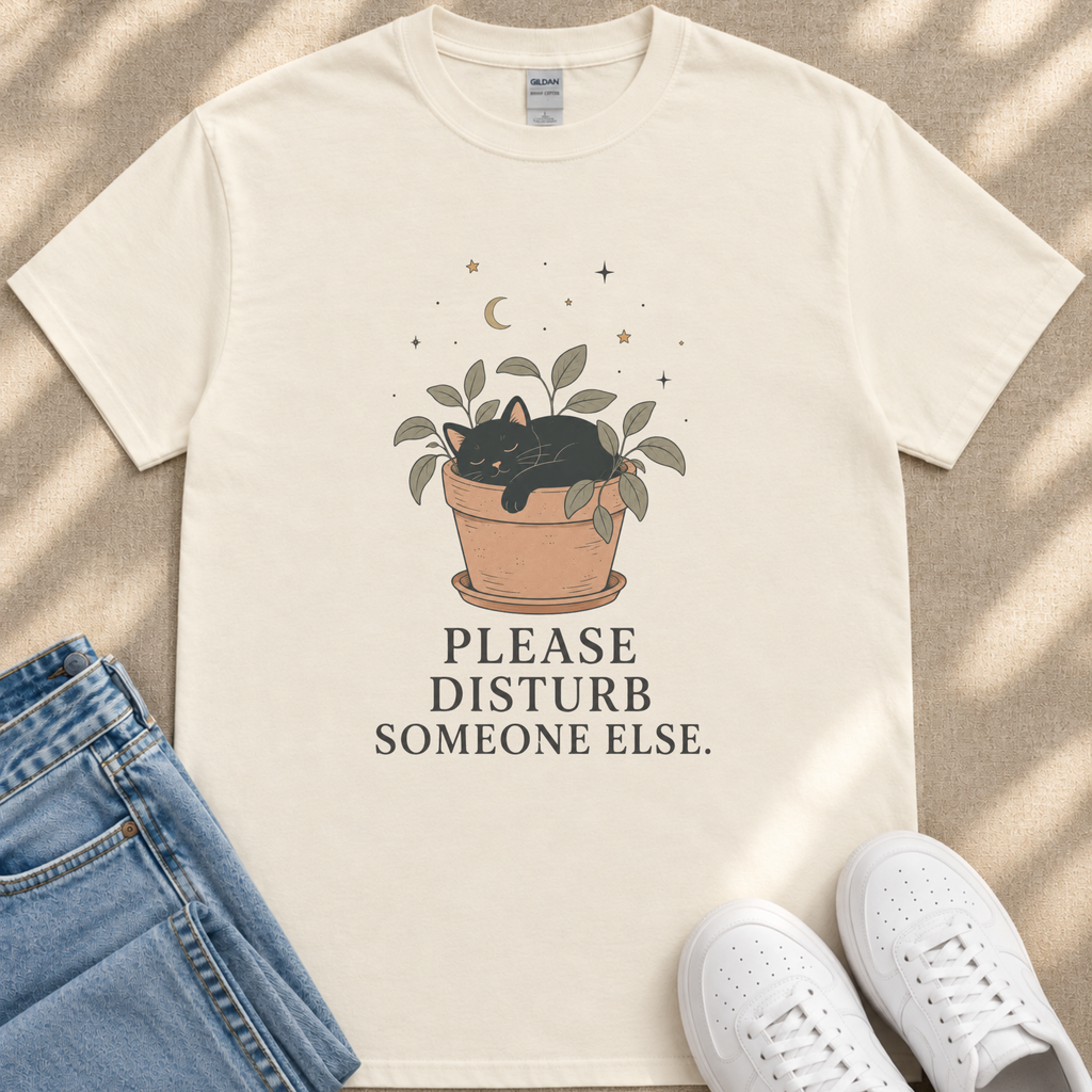 A white t-shirt with a graphic design of a cat sleeping in a pot, accompanied by the text "PLEASE DISTURB SOMEONE ELSE." The t-shirt is placed on a beige surface, with a pair of blue jeans and white sneakers visible in the background.