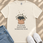 A white t-shirt with a graphic design of a cat sleeping in a pot, accompanied by the text "PLEASE DISTURB SOMEONE ELSE." The t-shirt is placed on a beige surface, with a pair of blue jeans and white sneakers visible in the background.