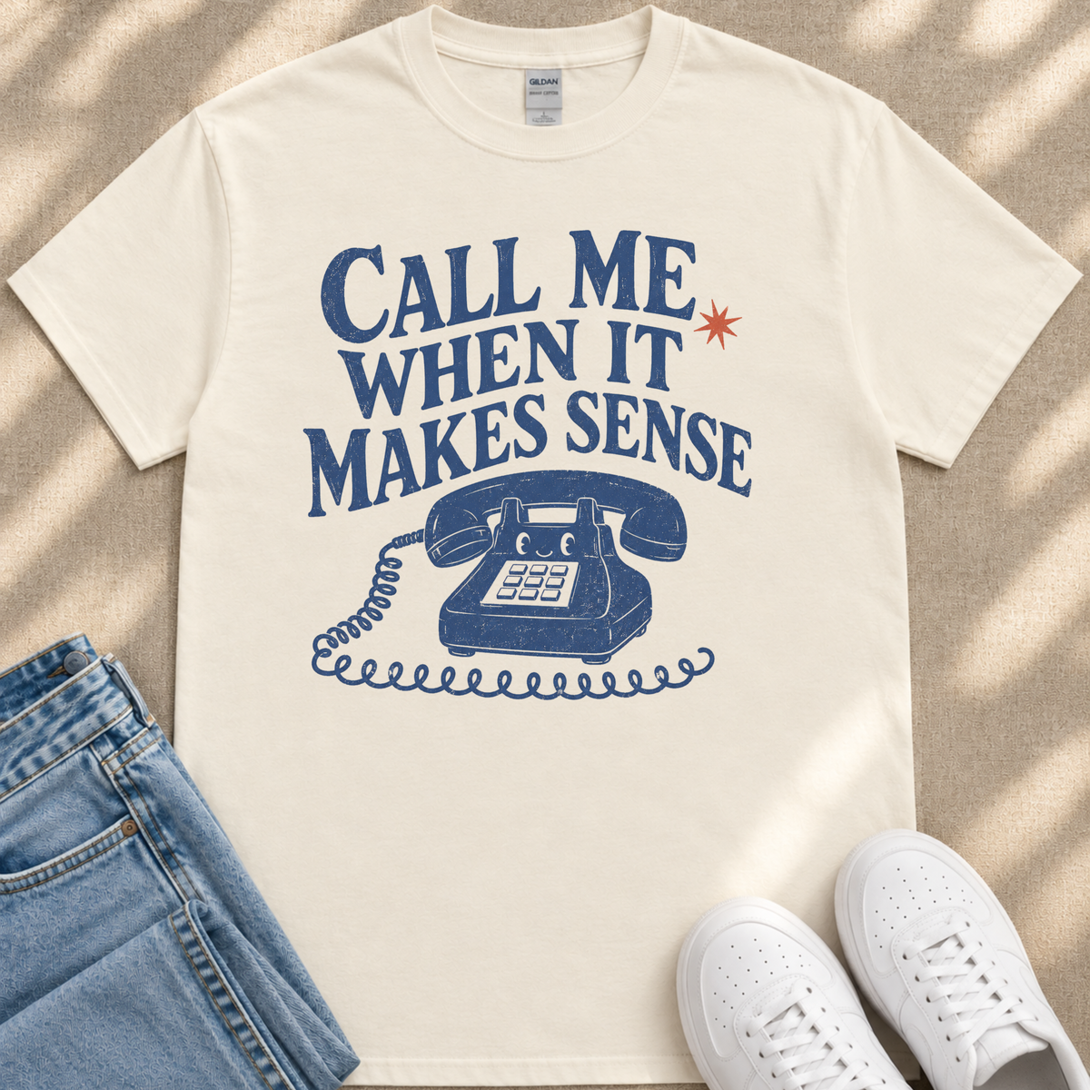 A white t-shirt with a graphic design of a telephone and the text "Call me when it makes sense" printed on it, along with a small star above the text. The t-shirt is placed next to a pair of blue jeans and white sneakers.