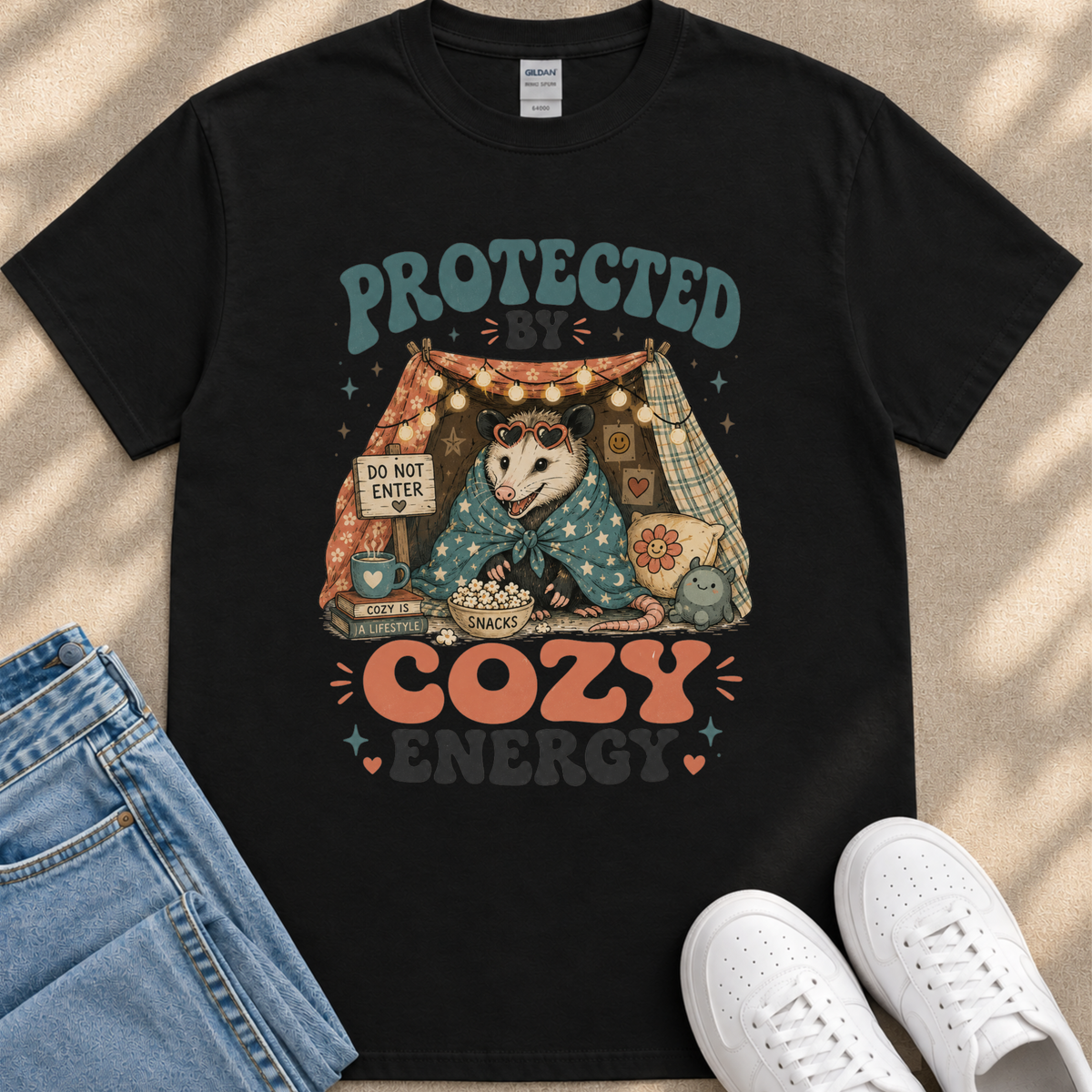 A black t-shirt with a graphic design featuring an opossum wrapped in a blanket, sitting inside a tent with a sign that says "Do not enter". The design also includes the text "Protected by Cozy Energy".