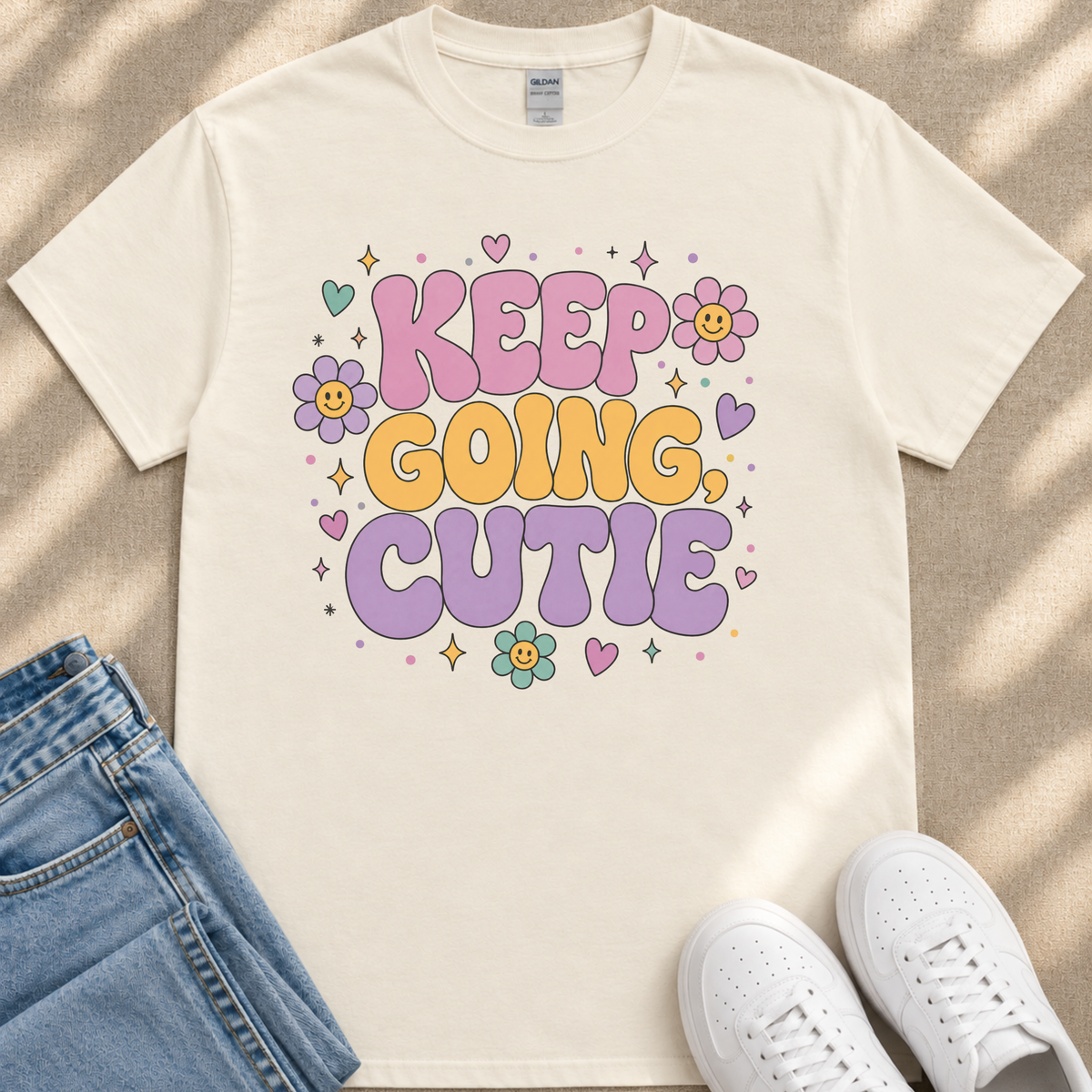A white t-shirt with a colorful graphic design that says "KEEP GOING CUTIE" surrounded by flowers and stars. The t-shirt is placed next to a pair of jeans and white sneakers.