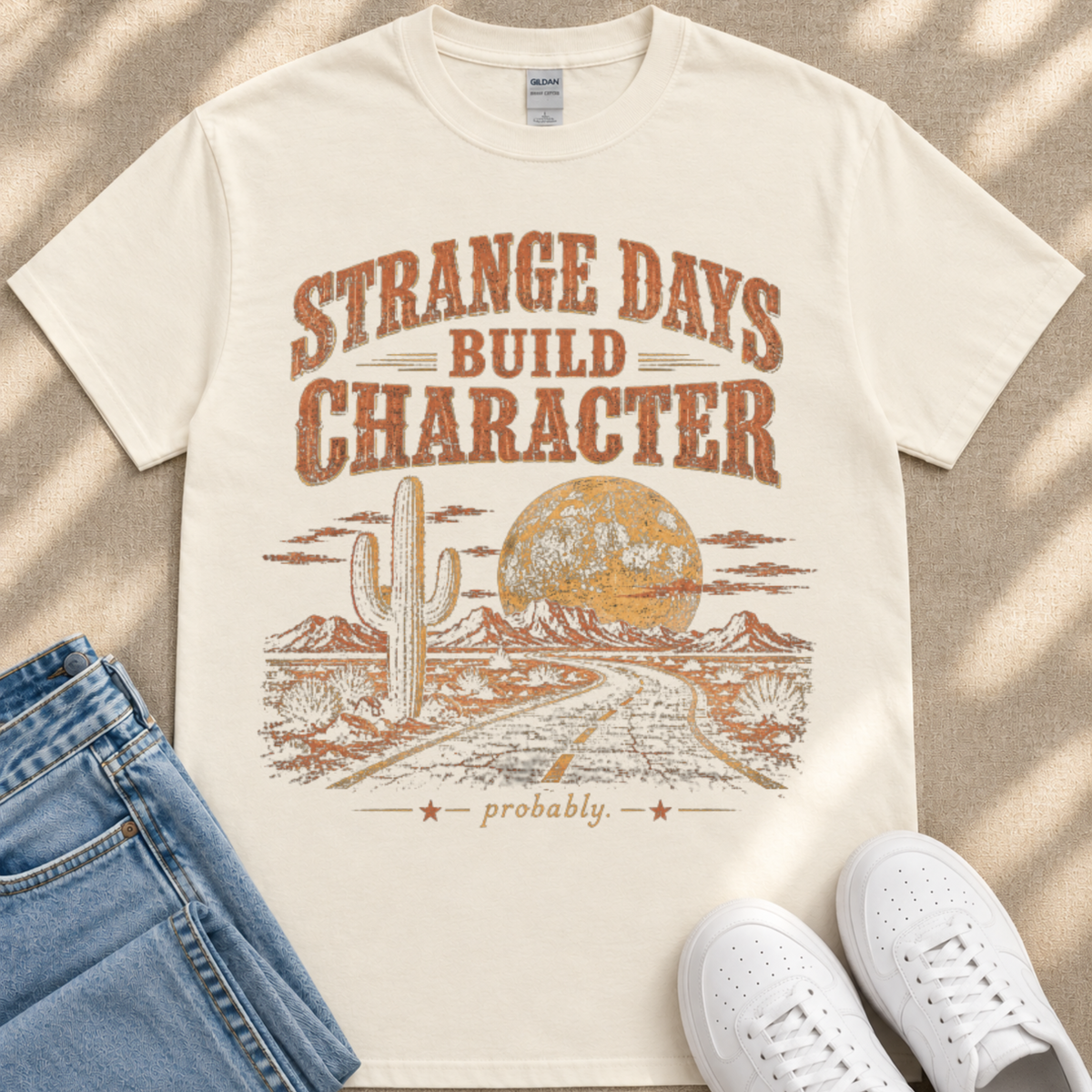 a white t-shirt with a graphic design depicting a desert landscape with a cactus and a road leading to a large moon. The text on the t-shirt reads " Strange Days Build Character probably".