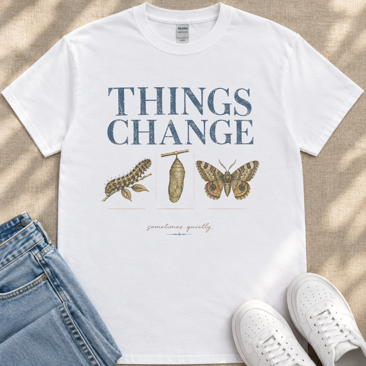 A white t-shirt with the text "THINGS CHANGE" printed on it, along with illustrations of a caterpillar and a butterfly. The t-shirt is placed on a beige surface, and there are a pair of jeans and white sneakers visible in the image.