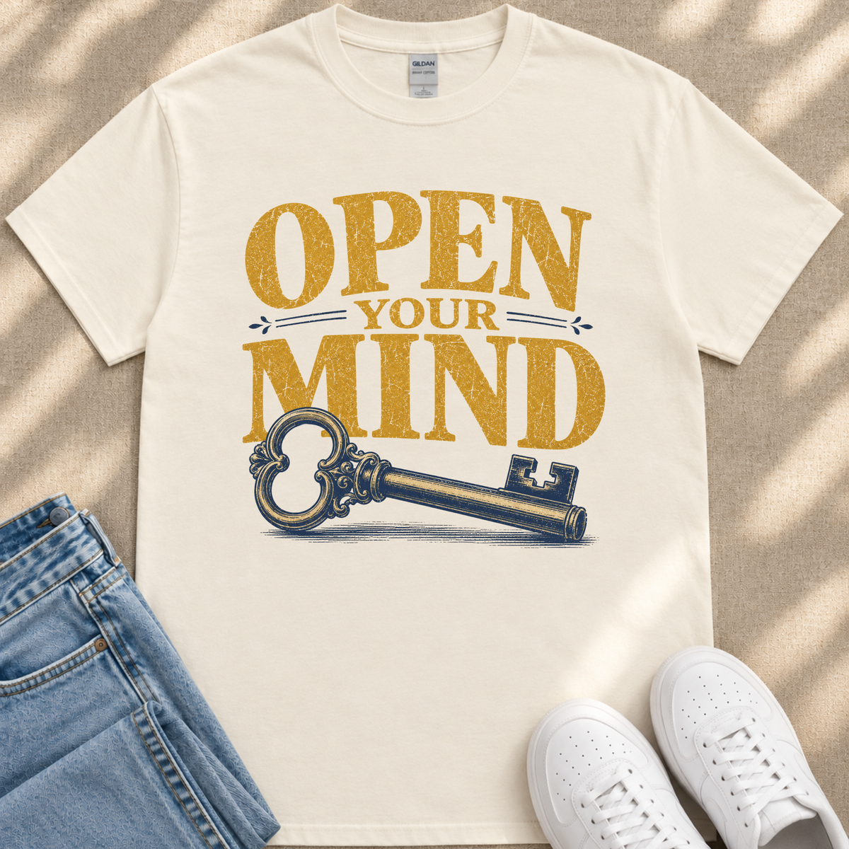 a white t-shirt with a graphic design of a key and the text "OPEN YOUR MIND" printed on it. The t-shirt is placed on a beige surface, and there are a pair of jeans and white sneakers visible in the image.