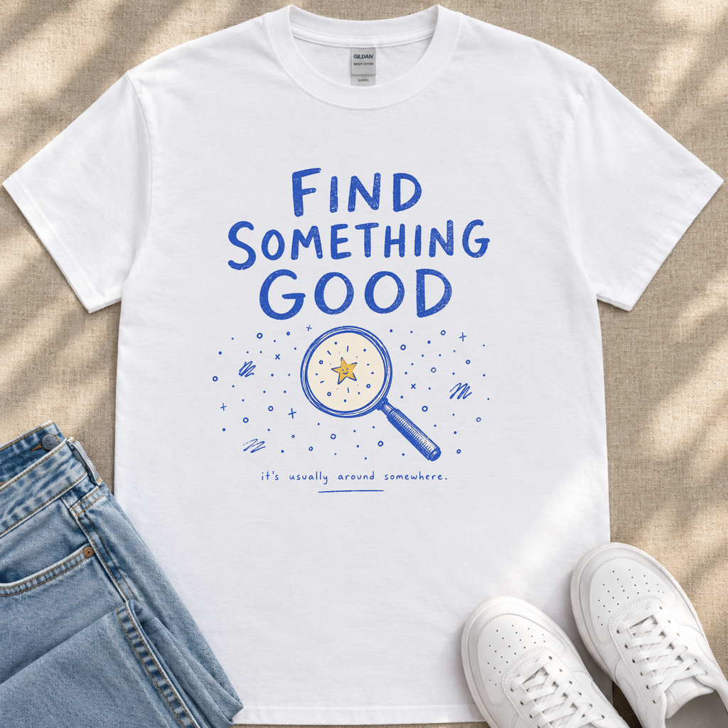 A white t-shirt with a blue graphic design and text, along with a pair of jeans and white sneakers, laid out on a beige surface.