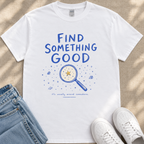 A white t-shirt with a blue graphic design and text, along with a pair of jeans and white sneakers, laid out on a beige surface.