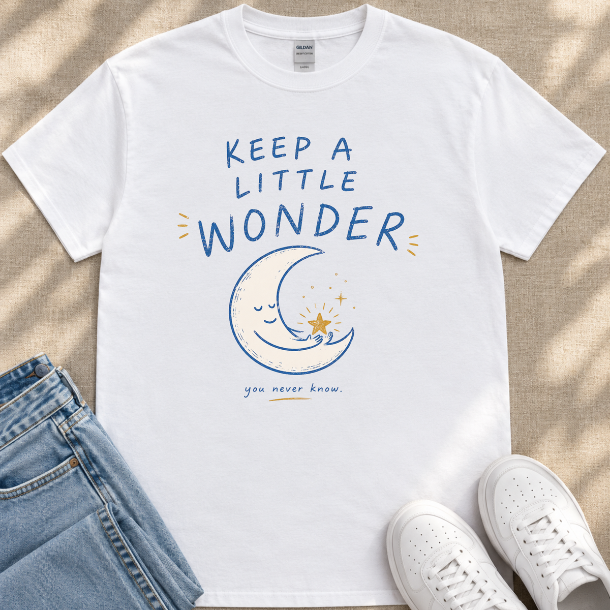 A white t-shirt with a graphic design of a crescent moon and stars, along with the text "KEEP A LITTLE WONDER" and a smaller text below that says "you never know." The t-shirt is placed on a beige surface, and there are a pair of blue jeans and white sneakers visible in the image.