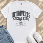 A white t-shirt with a graphic design that says "INTROVERT'S SOCIAL CLUB" and a chair graphic, along with the text "EST. 2023" and "ATTENDANCE OPTIONAL". The t-shirt is placed on a beige surface, and there are a pair of blue jeans and white sneakers visible in the image.