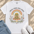 A white t-shirt with a colorful graphic design depicting a frog sitting on a mushroom, surrounded by flowers and a rainbow. The text on the t-shirt reads "Small steps still count" and "Even the wobbly ones".