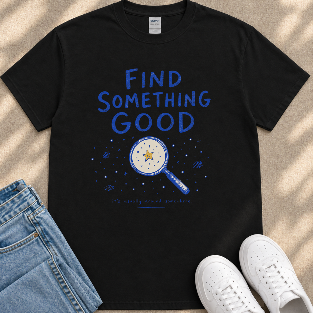 a black t-shirt with a blue design that includes a magnifying glass and stars, along with the text "Find something good" and "it's usually around somewhere." The t-shirt is placed on a beige surface, and there are a pair of blue jeans and white sneakers visible in the image.