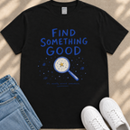 a black t-shirt with a blue design that includes a magnifying glass and stars, along with the text "Find something good" and "it's usually around somewhere." The t-shirt is placed on a beige surface, and there are a pair of blue jeans and white sneakers visible in the image.