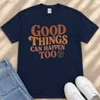 A navy blue t-shirt with the text "GOOD THINGS CAN HAPPEN TO" printed on it, along with a heart symbol. The t-shirt is placed on a beige surface, and there are a pair of jeans and white sneakers visible in the image.