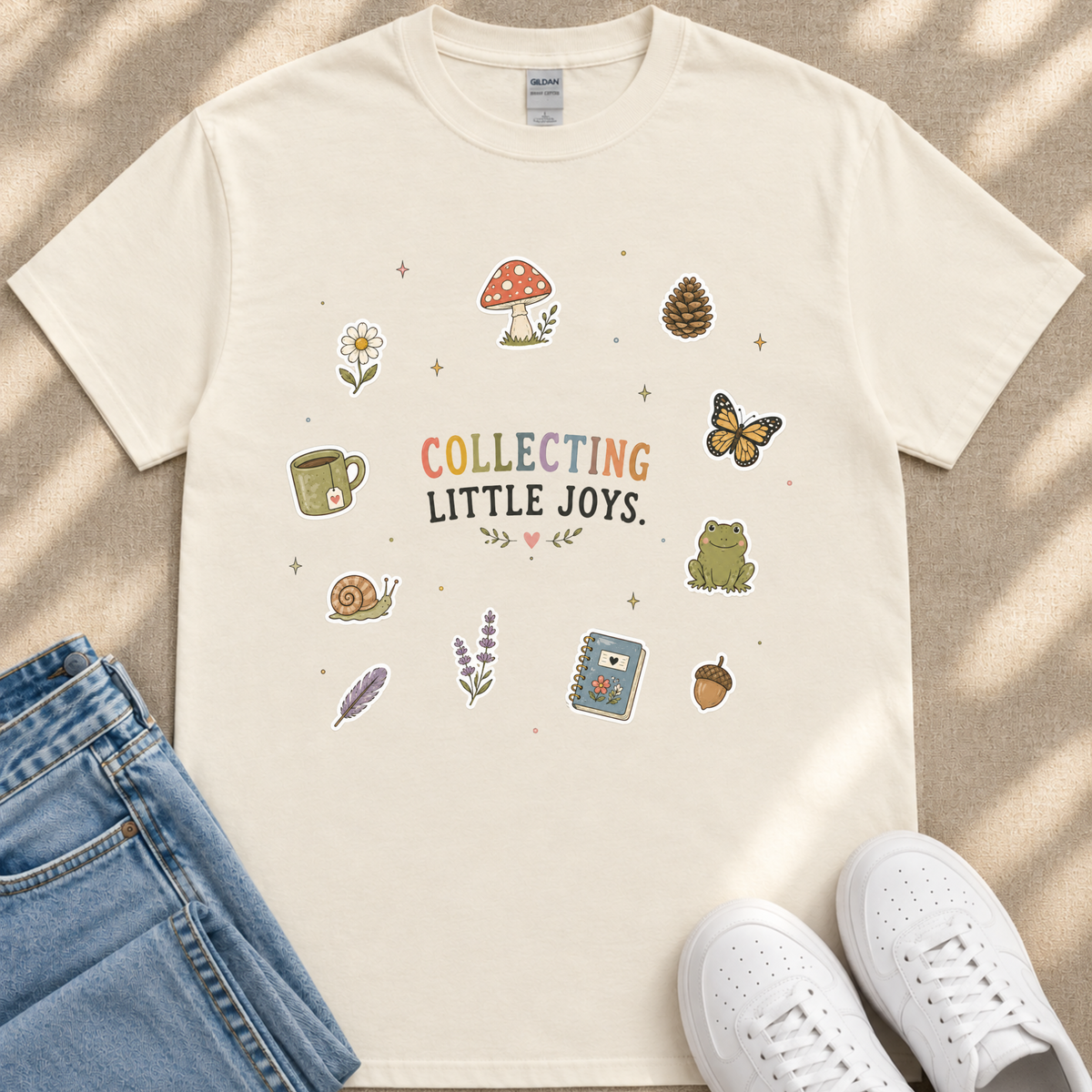 A white t-shirt with a colorful graphic design featuring various objects such as a mushroom, flower, butterfly, cup, frog, and more, along with the text "Collecting Little Joys". The t-shirt is placed on a beige surface, and there are a pair of jeans and white sneakers visible in the image.