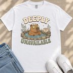 a white t-shirt with a graphic design depicting a brown bear sitting in a hot tub, surrounded by various elements such as a cup, a plant, and a pair of jeans. The text on the t-shirt reads "DEEPLY UNAVAILABLE".