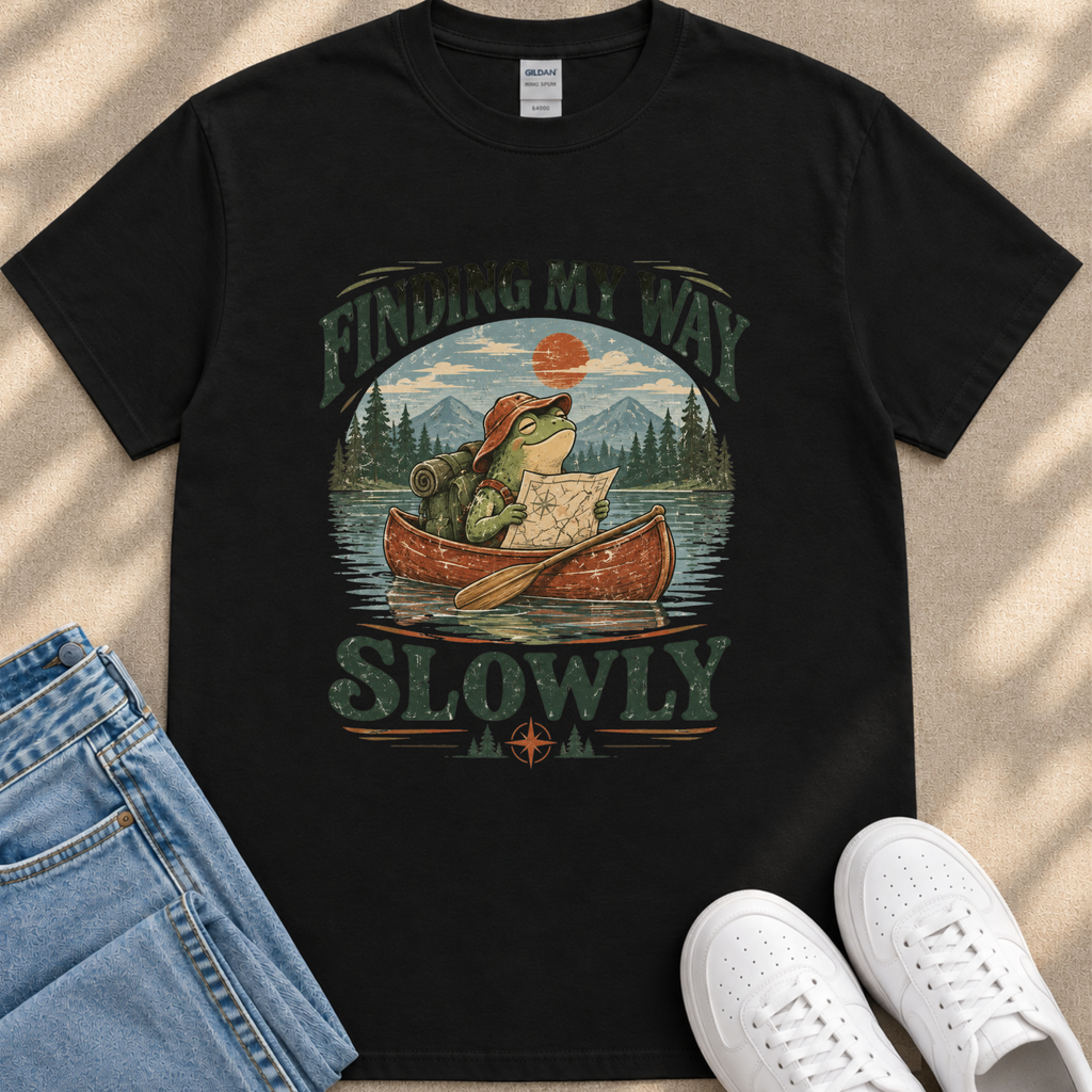 A black t-shirt with a graphic design depicting a frog in a canoe, holding a map and paddling slowly, with the text "FINDING MY WAY SLOWLY" printed on it.