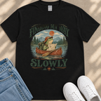 A black t-shirt with a graphic design depicting a frog in a canoe, holding a map and paddling slowly, with the text "FINDING MY WAY SLOWLY" printed on it.