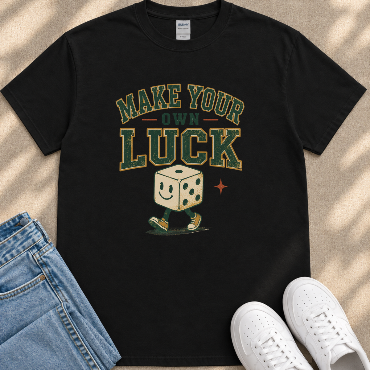 A black t-shirt with a graphic design featuring a smiling dice character and the text "MAKE YOUR OWN LUCK" printed on it.