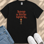 A black t-shirt with the text "keep a little spark" printed on it, along with an illustration of a lit match. The t-shirt is placed next to a pair of blue jeans and white sneakers.