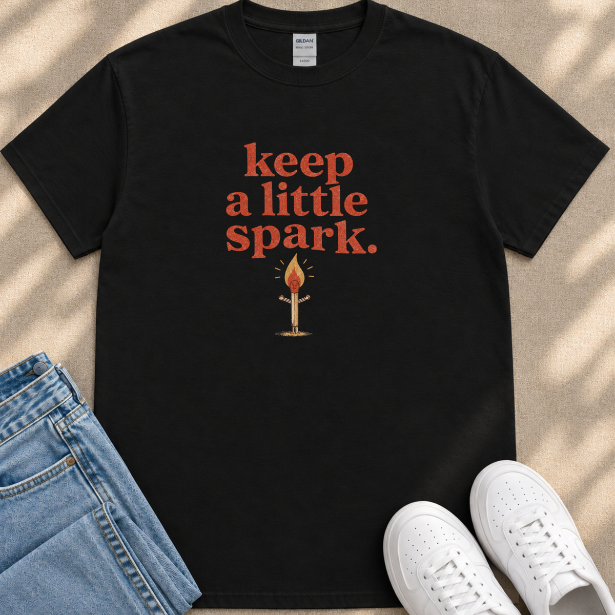 A black t-shirt with the text "keep a little spark" printed on it, along with an illustration of a lit match. The t-shirt is placed next to a pair of blue jeans and white sneakers.