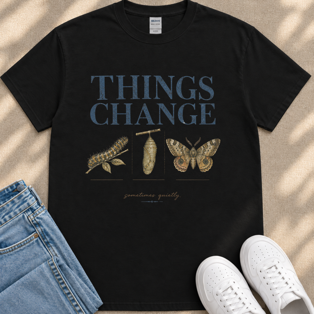 A black t-shirt with the text "THINGS CHANGE" printed on it, along with illustrations of a caterpillar and a butterfly. The t-shirt is placed on a beige surface, and there are a pair of jeans and white sneakers visible in the image.