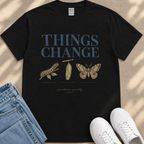 A black t-shirt with the text "THINGS CHANGE" printed on it, along with illustrations of a caterpillar and a butterfly. The t-shirt is placed on a beige surface, and there are a pair of jeans and white sneakers visible in the image.