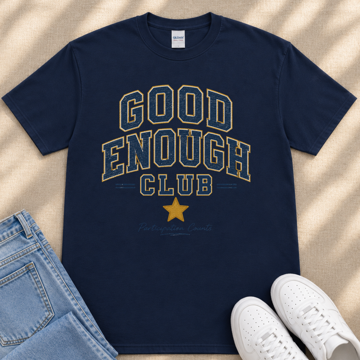 A navy blue t-shirt with the text "GOOD ENOUGH CLUB" printed on it, along with a yellow star symbol. The t-shirt is placed on a beige surface, and there are a pair of blue jeans and white sneakers visible in the image.
