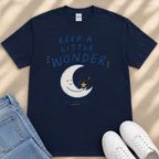 A navy blue t-shirt with a moon and star graphic, along with the text "KEEP A LITTLE WONDER" and a smaller text below that reads "you never know." The t-shirt is laid out on a beige surface, and there are a pair of jeans and white sneakers visible in the image.