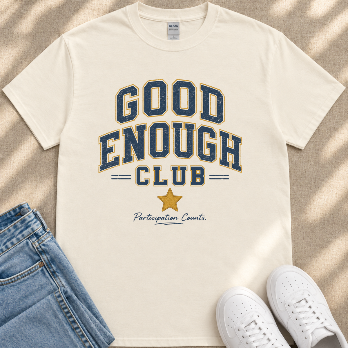 A white t-shirt with the text "GOOD ENOUGH CLUB Participation Counts" printed on it, along with a yellow star symbol. The t-shirt is placed on a beige surface, and there are a pair of blue jeans and white sneakers visible in the image.