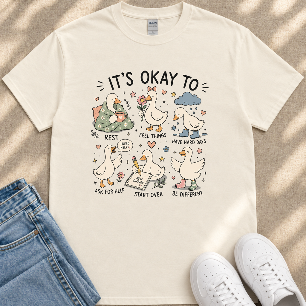 A white t-shirt with a graphic design featuring cartoon ducks and text that says "It's OKAY TO".