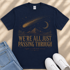 A navy blue t-shirt with a graphic design depicting a shooting star, mountains, and a crescent moon, along with the text "WE'RE ALL JUST PASSING THROUGH" and "MIGHT AS WELL LOOK AROUND" printed on it.