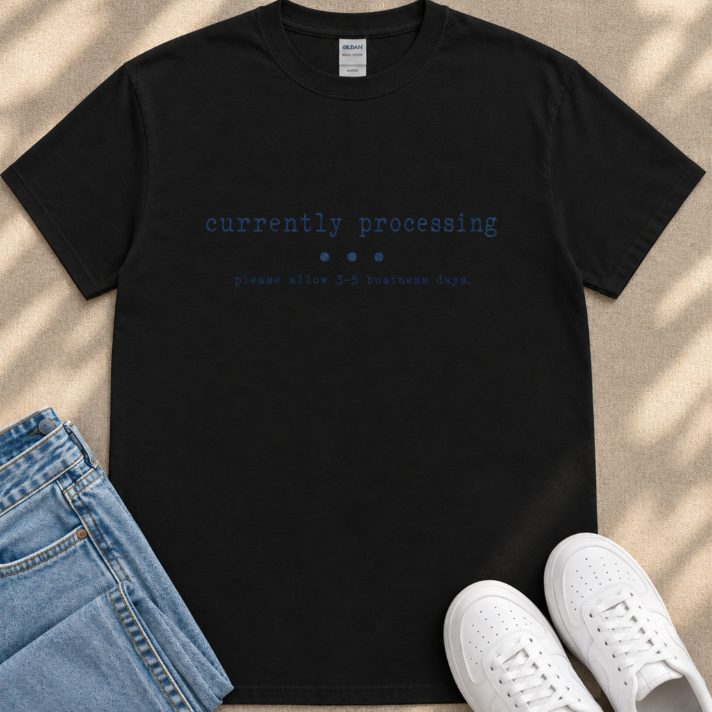 a black t-shirt with the text "currently processing" printed on it, along with instructions to allow 3-5 business days. The t-shirt is placed on a beige surface, and there are a pair of blue jeans and white sneakers visible in the image.