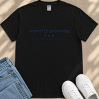 a black t-shirt with the text "currently processing" printed on it, along with instructions to allow 3-5 business days. The t-shirt is placed on a beige surface, and there are a pair of blue jeans and white sneakers visible in the image.