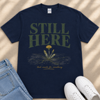 A navy blue t-shirt with the text "STILL HERE" printed on it, along with an illustration of a dandelion flower. The t-shirt is laid out on a beige surface, and there are a pair of jeans and white sneakers visible in the image.